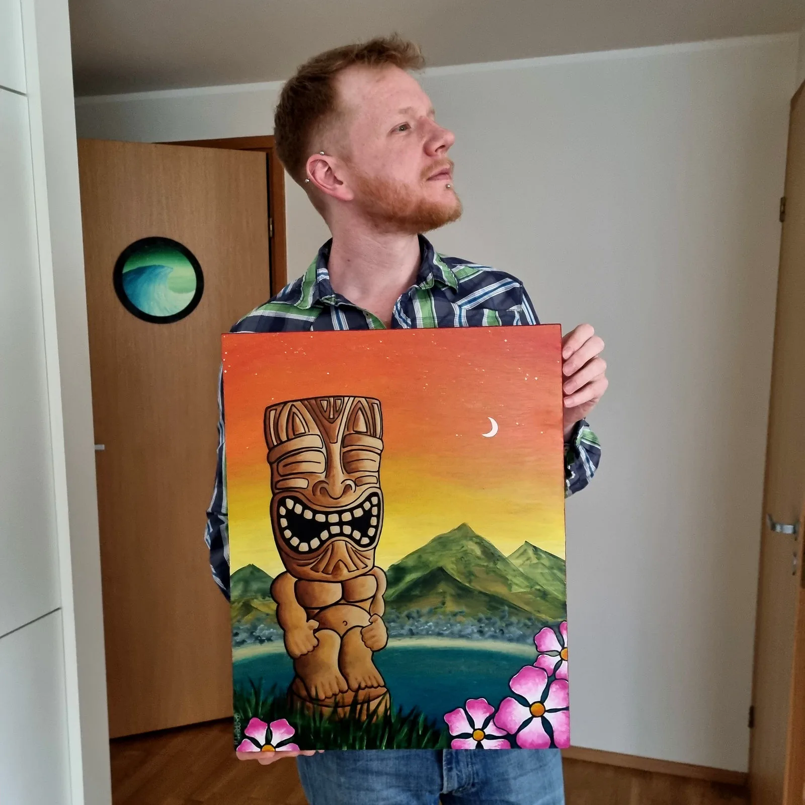 Holding a painting of a smiling tiki statue in front of mountains and a sunset