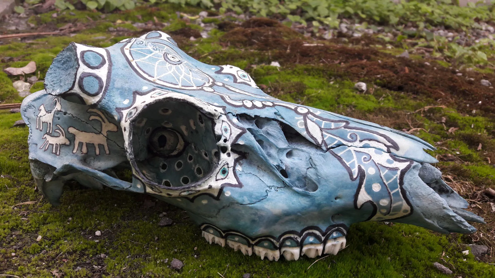 Animal skull painted pale blue with silver patterns, resting on moss